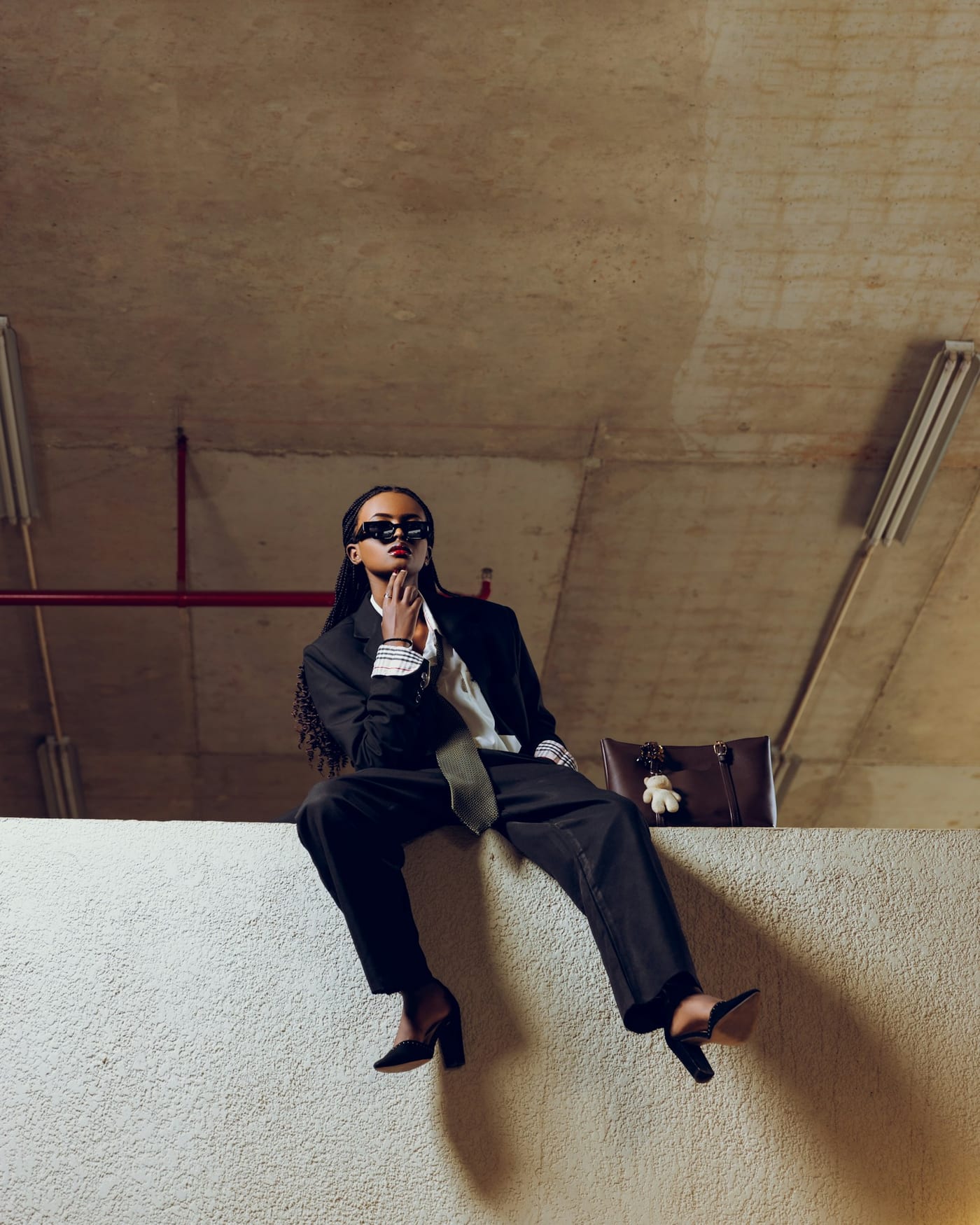 Woman in sunglasses, a black oversized suit, striped shirt cuffs, a patterned tie and black heels, sitting on a concrete ledge
