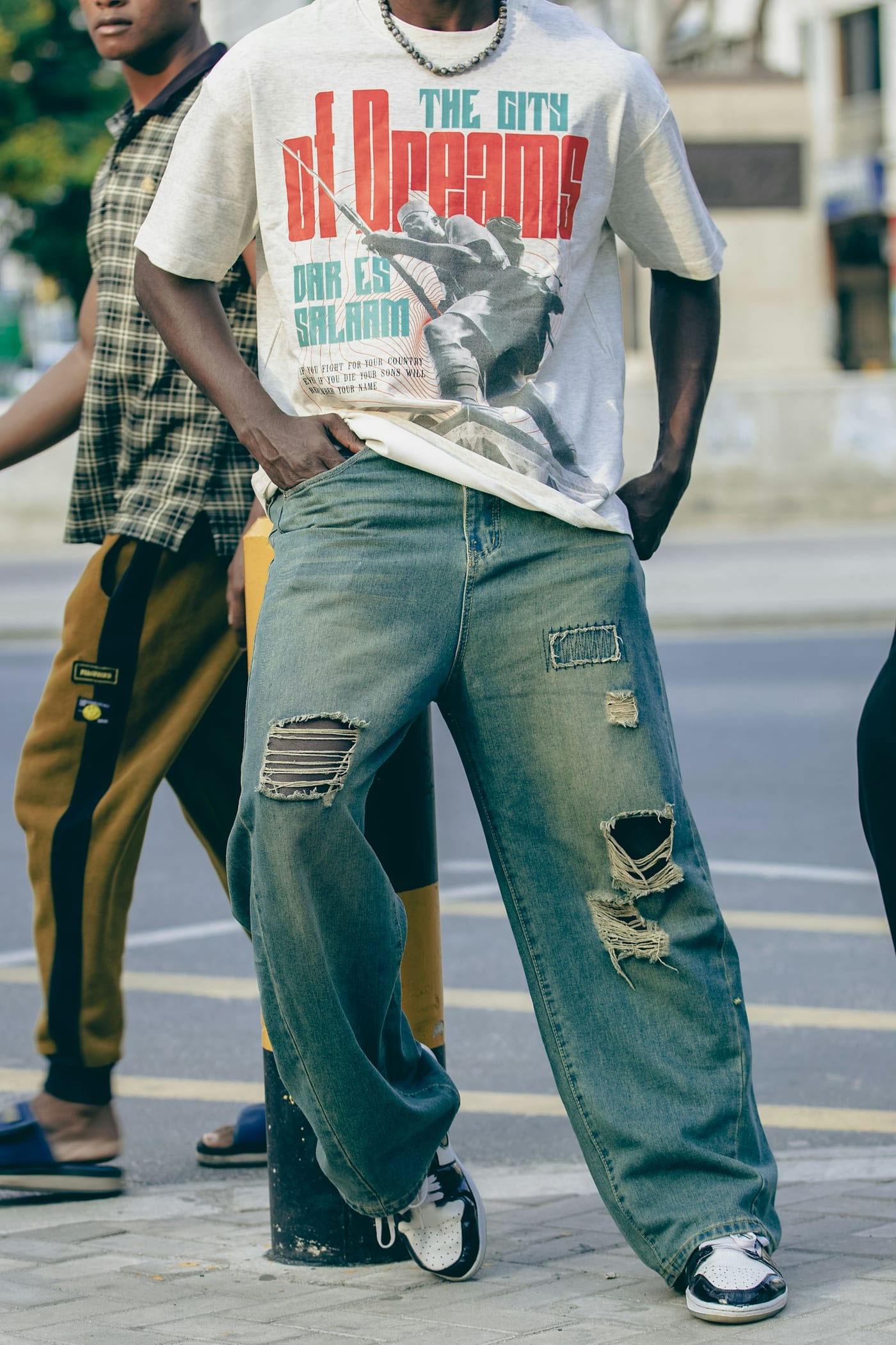 Man in a graphic tee, a bead necklace, wide ripped jeans and two-tone trainers on a city street