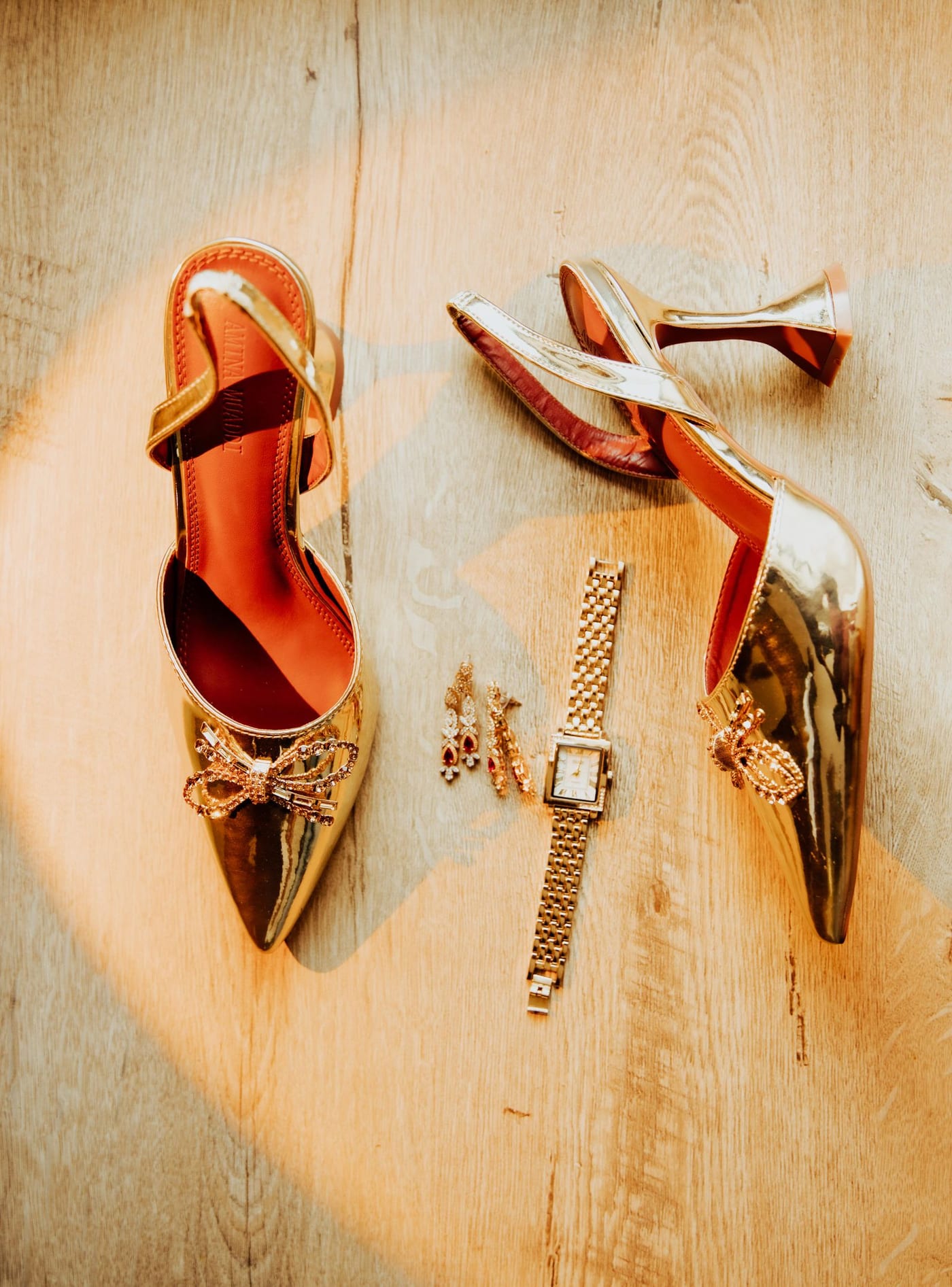Gold metallic slingback heels with crystal bows, drop earrings and a gold watch laid on a wooden floor