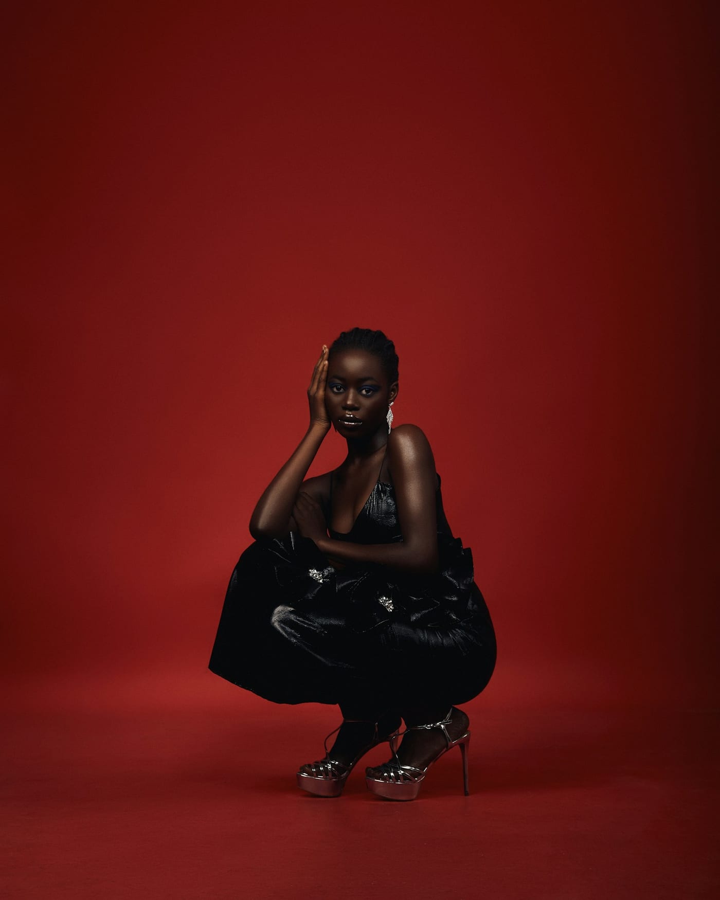 Woman in a black satin dress and silver platform sandals, crouching against a red backdrop